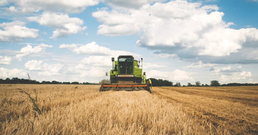 A green tractor harvesting wheat in a vast open field under a bright, cloudy sky.